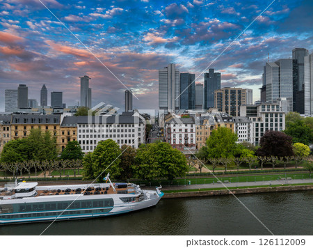 Aerial view of Frankfurt, Germany, featuring the Commerzbank Tower, modern skyscrapers, the Main River with a docked riverboat, and a vibrant sunset sky. Aerial view of Frankfurt, Germany, featuring the Commerzbank Tower, modern skyscrapers, the Main River with a docked riverboat, and a vibrant sunset sky. 126112009