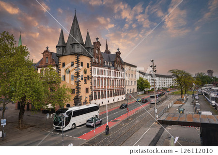 A street in Frankfurt, Germany, featuring the historic Saalhof building with pointed towers, a tree lined road, a red bike lane, and moored boats by the riverside. 126112010