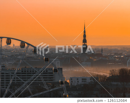 Serene sunrise over Riga, Latvia, featuring St. Peter's Church spire, a Ferris wheel in the foreground, and a warm orange sky with soft morning light. 126112084