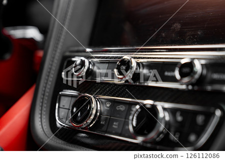 Detailed view of a luxury car's control panel with chrome knobs, carbon fiber trim, and red leather interior, showcasing premium craftsmanship. 126112086