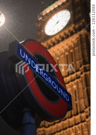 A London Underground sign in red and blue stands in the foreground, with Big Ben's illuminated clock face visible at night under a faint drizzle. 126112088