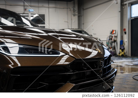 Front end of a metallic brown Lamborghini Urus with sharp LED headlight and emblem, set in a garage with tools, hose, and cleaning supplies visible. 126112092
