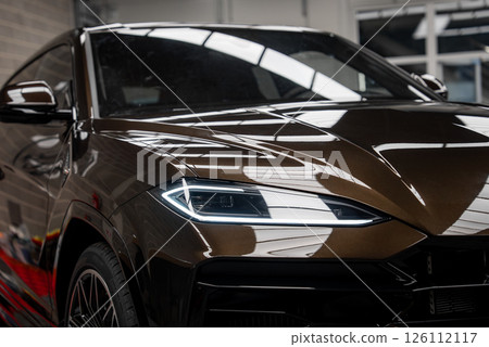 Close up view of a Lamborghini Urus showcasing angular headlights, glossy brown exterior, and reflections of overhead lights in an indoor showroom. 126112117
