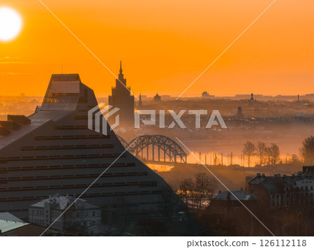 A sunrise view of Riga, Latvia, showcasing the National Library, Riga TV Tower, Daugava River, and arched railway bridge under a misty orange sky. 126112118