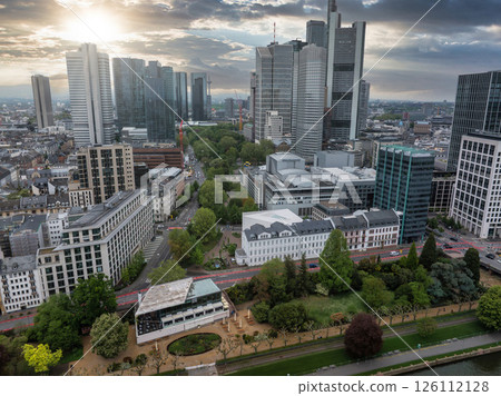 Aerial view of Frankfurt, Germany, featuring modern skyscrapers like Commerzbank Tower, a green park, the Main River, and a dramatic sunset sky. 126112128