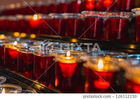 Red votive candles in glass holders, some lit and others unlit, arranged on a black metal stand in a serene religious or spiritual setting. Red votive candles in glass holders, some lit and others unlit, arranged on a black metal stand in a serene religious or spiritual setting. 126112150