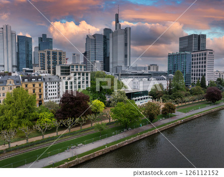 Aerial view of Frankfurt, Germany, featuring the Commerzbank Tower, riverside park greenery, and vibrant orange and pink sunset skies over the Main River. 126112152