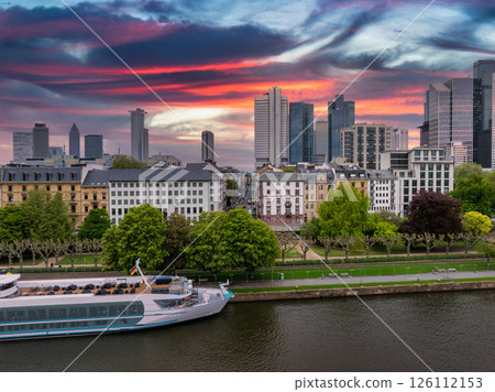 Aerial view of Frankfurt, Germany, at sunset featuring modern skyscrapers like Commerzbank Tower, the Main River, and a white riverboat by a tree lined promenade. 126112153