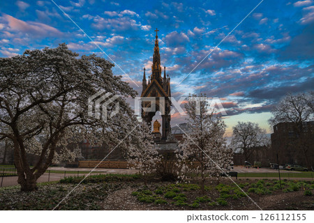 The Albert Memorial in London stands amidst blooming white trees, featuring a golden statue of Prince Albert under a vibrant blue sky with scattered clouds. 126112155