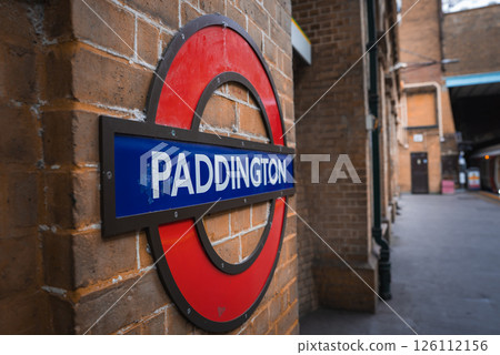 The iconic red and blue Paddington roundel sign on a brick wall, with a train partially visible on the platform of the London Underground station. 126112156