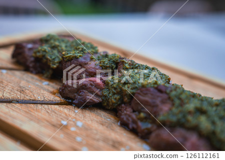 Slices of cooked meat topped with green herb based sauce on a wooden cutting board. The background shows blurred greenery, suggesting an outdoor setting. 126112161