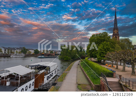 View of Frankfurt, Germany, featuring the Main River, a docked boat, Dreikonigskirche spire, a tree lined pathway, and a mix of urban architecture. View of Frankfurt, Germany, featuring the Main River, a docked boat, Dreikonigskirche spire, a tree lined pathway, and a mix of urban architecture. 126112169