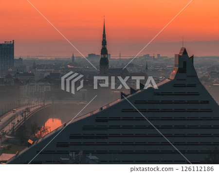 Serene sunrise over Riga, Latvia, featuring St. Peter's Church spire, historic cityscape, modern Latvian National Library, bridge, and waterway. Serene sunrise over Riga, Latvia, featuring St. Peter's Church spire, historic cityscape, modern Latvian National Library, bridge, and waterway. 126112186