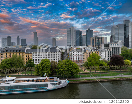 Aerial view of Frankfurt, Germany, featuring the skyline with Commerzbank Tower, the Main River, a docked cruise ship, and a vibrant sunset sky. Aerial view of Frankfurt, Germany, featuring the skyline with Commerzbank Tower, the Main River, a docked cruise ship, and a vibrant sunset sky. 126112200