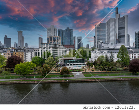 Aerial view of Frankfurt, Germany, featuring the skyline with Commerzbank Tower, riverside park along the Main River, and a vibrant sunset sky. 126112223