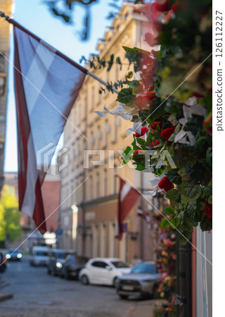 A cobblestone street in Riga's old town features a Latvian flag, pastel colored buildings, parked cars, and vibrant flowers under a clear blue sky. A cobblestone street in Riga's old town features a Latvian flag, pastel colored buildings, parked cars, and vibrant flowers under a clear blue sky. 126112227