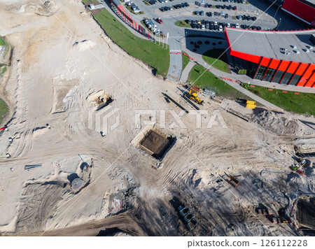 Aerial view of a construction site in Riga, Latvia, featuring sandy terrain, a large excavation pit, machinery, and a nearby modern red building. 126112228