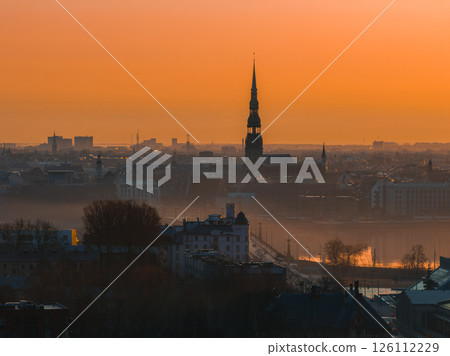 Serene sunrise over Riga, Latvia, featuring St. Peter's Church spire, the Daugava River, historic rooftops, and a light mist enhancing the scene. Serene sunrise over Riga, Latvia, featuring St. Peter's Church spire, the Daugava River, historic rooftops, and a light mist enhancing the scene. 126112229