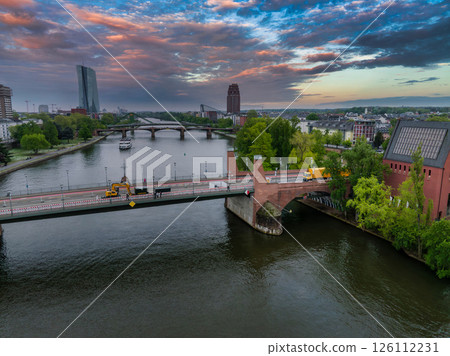 Aerial image of Frankfurt, Germany, showing the Main River, a bridge with construction vehicles, the ECB building, and a red brick tower at sunset. Aerial image of Frankfurt, Germany, showing the Main River, a bridge with construction vehicles, the ECB building, and a red brick tower at sunset. 126112231