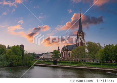The Frankfurt Cathedral's Gothic spire rises near the Main River, surrounded by greenery, a riverside walkway, and urban architecture under an overcast sky. The Frankfurt Cathedral's Gothic spire rises near the Main River, surrounded by greenery, a riverside walkway, and urban architecture under an overcast sky. 126112235