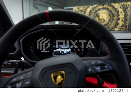 Close up of a Lamborghini interior showing the steering wheel with logo, digital dashboard displaying a Urus, and red accents in a luxury showroom. 126112244