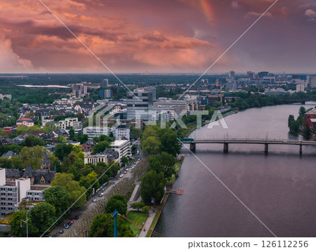 Aerial image of Frankfurt, Germany, featuring the Main River, a bridge, urban skyline, lush greenery, and a vibrant sunset with a faint rainbow. 126112256