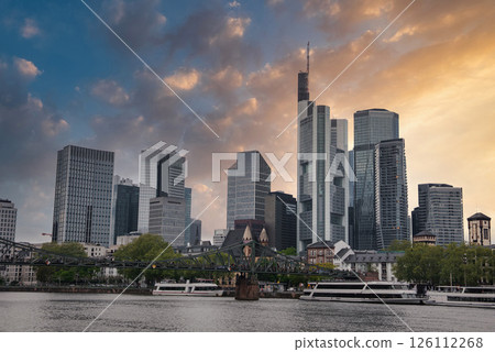 Frankfurt skyline featuring modern skyscrapers, the Commerzbank Tower, the Main River, Eiserner Steg, historic church spire, trees, and boats. 126112268