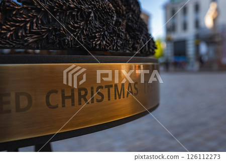 A metallic plaque with engraved text and intricate pine branch metalwork, set against a cobblestone street and blurred buildings in Riga's old town. A metallic plaque with engraved text and intricate pine branch metalwork, set against a cobblestone street and blurred buildings in Riga's old town. 126112273