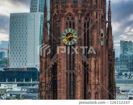 The Frankfurt Cathedral's Gothic architecture and red sandstone tower stand out against modern skyscrapers under a cloudy sky in Frankfurt, Germany. 126112287