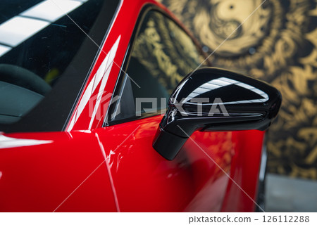 Side mirror and door of a red 2025 Porsche 911 GT3 922 with glossy black accents. Reflections and a golden abstract mural enhance the modern design. 126112288