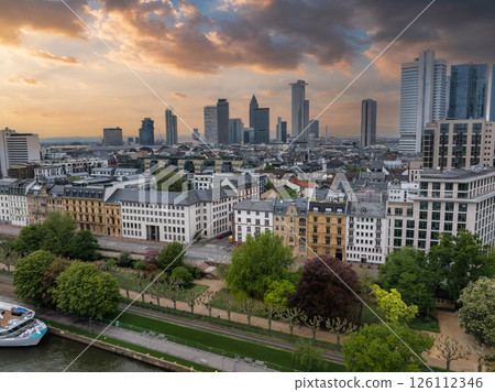 Aerial view of Frankfurt, Germany, featuring modern skyscrapers like Commerzbank Tower, a riverside promenade, a docked boat, and a dramatic sunset sky. 126112346