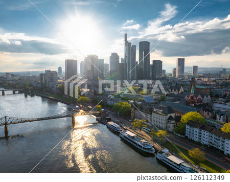Aerial view of Frankfurt, Germany, showcasing the skyline with Commerzbank Tower, the River Main, Eiserner Steg, and warm sunset lighting. 126112349