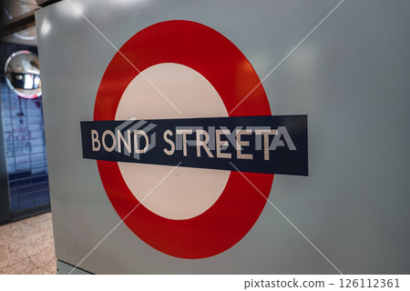 The iconic Bond Street Underground roundel sign with a red circle, blue bar, and white center, set against a light blue wall and tiled flooring. 126112361