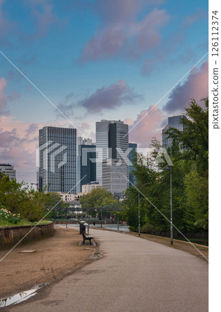 A park pathway with greenery and a bench leads to modern skyscrapers in Frankfurt, Germany. The overcast sky highlights the urban skyline. 126112374