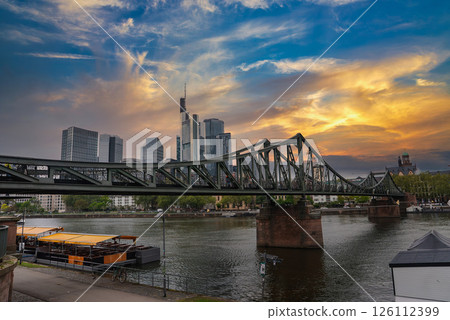 Eiserner Steg spans the Main River in Frankfurt, Germany, with a docked boat, historic cathedral spire, and modern skyscrapers under an overcast sky. 126112399