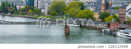 The Eiserner Steg pedestrian bridge spans the Main River in Frankfurt, Germany, with people crossing. Modern and historic architecture, including a church with twin spires, is visible in the The Eiserner Steg pedestrian bridge spans the Main River in Frankfurt, Germany, with people crossing. Modern and historic architecture, including a church with twin spires, is visible in the 126112409