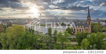 Aerial view of Frankfurt, Germany, featuring Dreikonigskirche's tall spire, lush green trees, mixed architecture, and a skyline at sunset with scattered clouds. Aerial view of Frankfurt, Germany, featuring Dreikonigskirche's tall spire, lush green trees, mixed architecture, and a skyline at sunset with scattered clouds. 126112410
