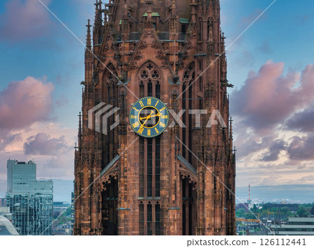 The Gothic style Frankfurt Cathedral tower stands out with intricate details and a gold accented clock, set against modern buildings and a pink blue sky. 126112441