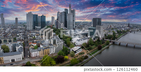 Aerial view of Frankfurt, Germany, featuring the skyline with Commerzbank Tower, the Main River, a bridge, and a vibrant sunset sky with pink and orange hues. 126112460