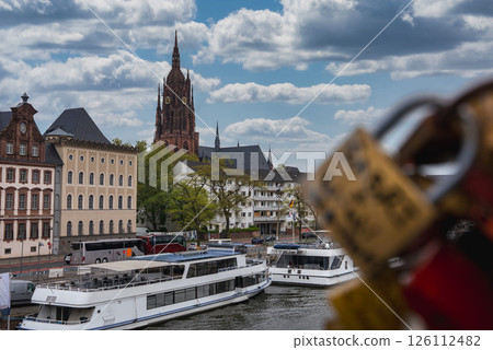 Frankfurt Cathedral rises in the background, with boats along the river, historic European buildings, and love locks on a bridge in the foreground. 126112482