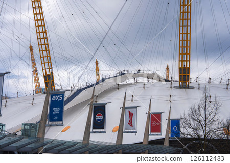 The O2 Arena in London features climbers on its white dome with yellow masts, brand banners in the foreground, and an overcast sky in the background. 126112483