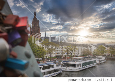 View of Frankfurt, Germany, featuring the Gothic style Frankfurt Cathedral, a bridge with love locks, the Main River, and docked riverboats. 126112501