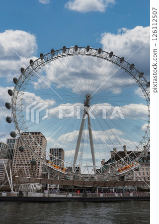The London Eye stands along the River Thames, framed by a partly cloudy sky. Modern and historic buildings surround the landmark, reflecting in the river. 126112507