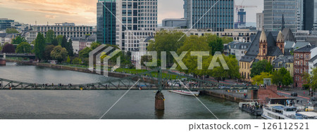 Aerial view of Frankfurt, Germany, showing the Eiserner Steg bridge over the Main River, modern skyscrapers, historic architecture, and a sunset sky. 126112521