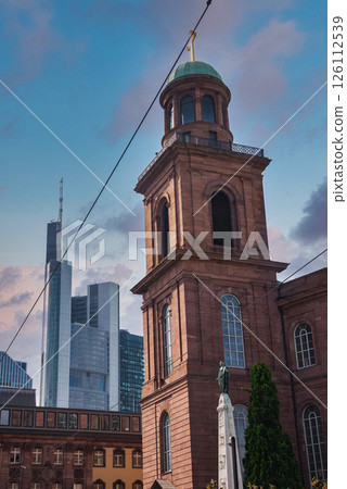 St. Paul's Church with its green dome and golden cross stands amid modern skyscrapers, including Commerzbank Tower, under a clear blue sky. 126112539
