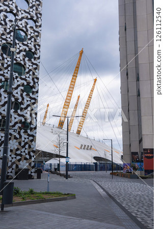 The O2 Arena's white dome and yellow masts stand out amid modern buildings, including one with a geometric facade, under a cloudy London sky. The O2 Arena's white dome and yellow masts stand out amid modern buildings, including one with a geometric facade, under a cloudy London sky. 126112540