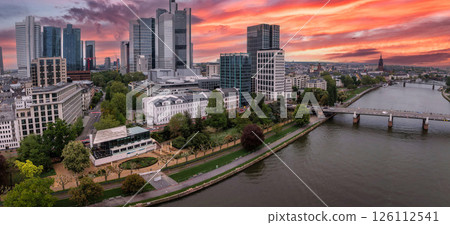 Aerial view of Frankfurt, Germany, at sunset featuring the Commerzbank Tower, Main River, a bridge, riverside greenery, and Frankfurt Cathedral spire. 126112541