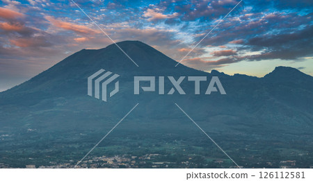 Mount Vesuvius rises with its conical shape, surrounded by greenery and a small urban area. The sky displays hues of blue, orange, and pink. 126112581