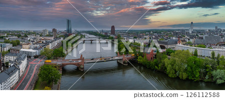 Aerial view of Frankfurt, Germany, featuring the Main River, European Central Bank, bridges, lush greenery, and a dramatic orange and purple sunset. Aerial view of Frankfurt, Germany, featuring the Main River, European Central Bank, bridges, lush greenery, and a dramatic orange and purple sunset. 126112588