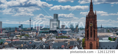 Aerial view of Frankfurt, Germany, highlighting the Gothic Frankfurt Cathedral, Europaturm, modern high rises, and rolling hills under a cloudy sky. 126112595